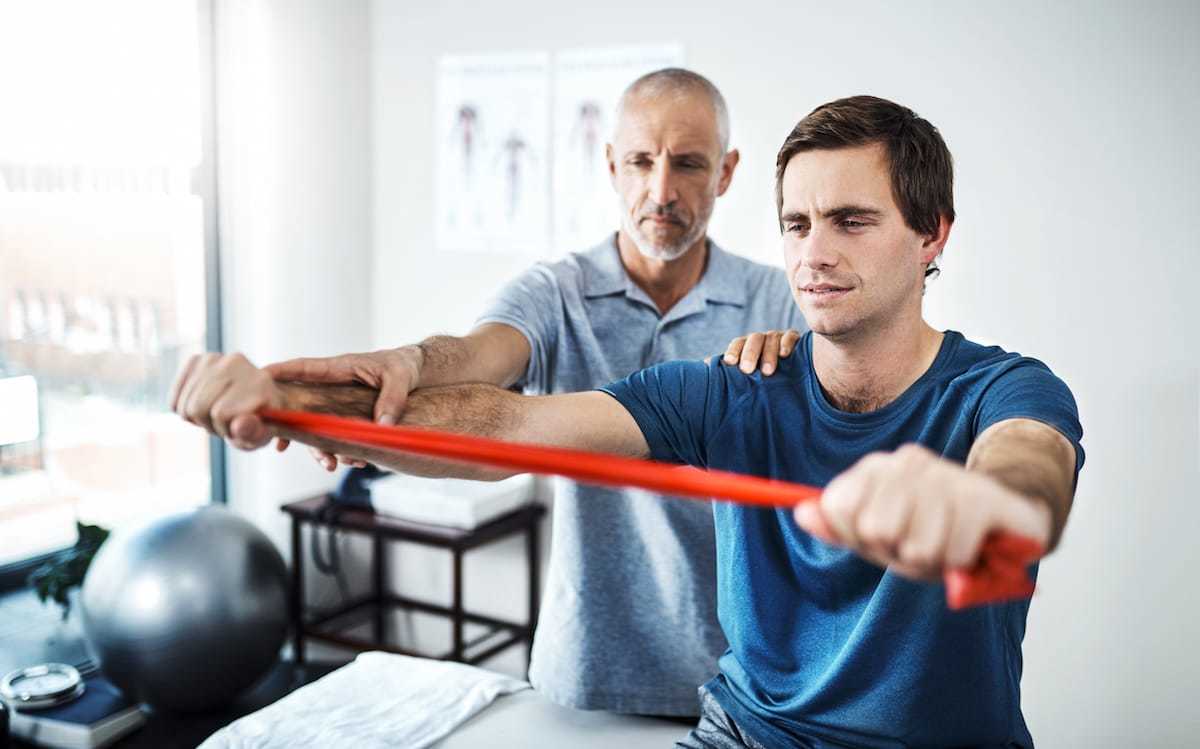 Ratgeber Rheuma: Ein Junder Mann fürht auf Anweisung eines Physiotrainers eine Halteübung mit einer Stange durch