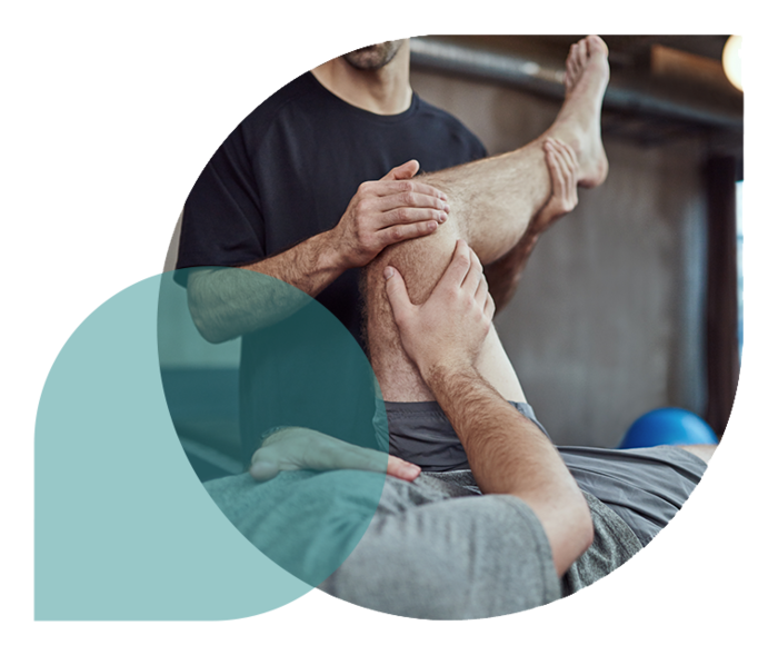 Ein Mann liegt auf dem Rücken und winkelt sein linkes Bein an, während ein anderer Mann seinen Unterschenkel mit den Händen hält.