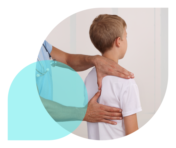 Ein Arzt tastet mit beiden Händen die rechte Schulter eines mit dem Rücken zu ihm stehenden Jungen ab