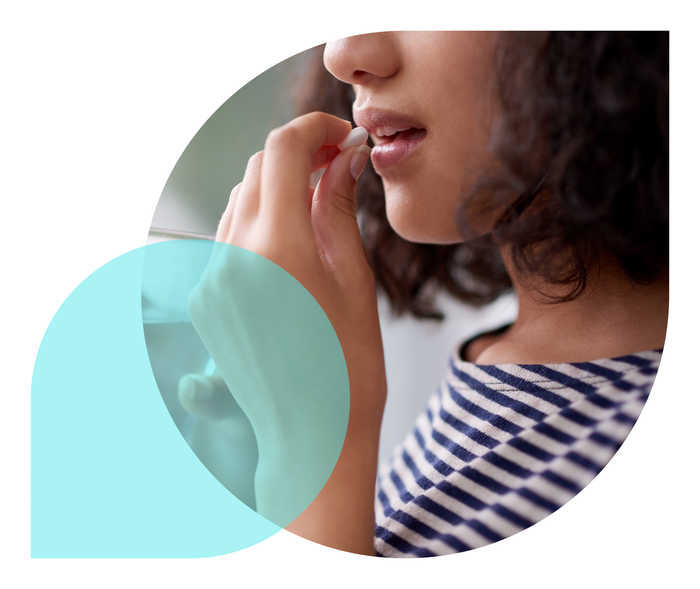 Eine Frau mit einem Glas Wasser in der rechten Hand führt mit der linken Hand eine Tablette zu ihrem Mund