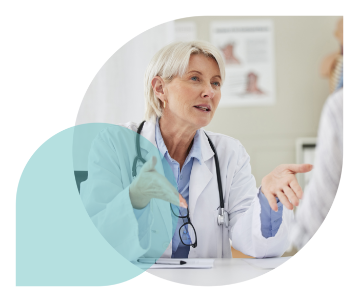 Eine Frau in einem weißen Kittel und mit einem um den Hals hängenden Stethoskop unterhält sich mit einer weiteren Frau.