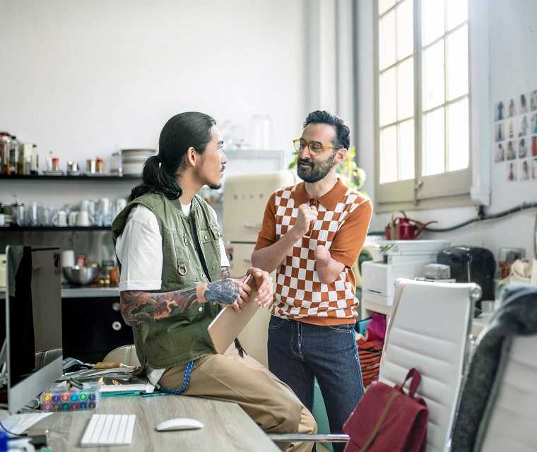 Morbus-Bechterew Patient Hun-Jong und mit seinem Kollegen Mesut unterhalten sich am Schreibtisch