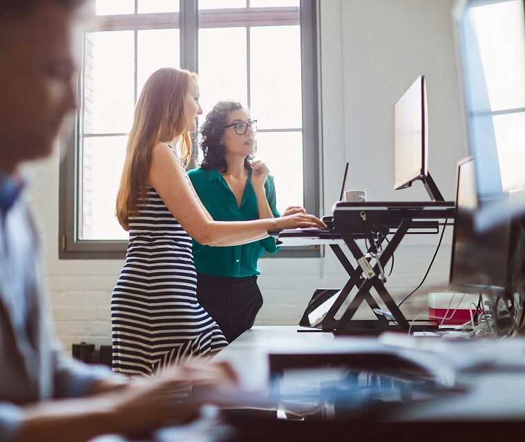 Zwei Kolleginnen stehen an ihrem Schreibtisch und schauen etwas auf dem Bildschirm an