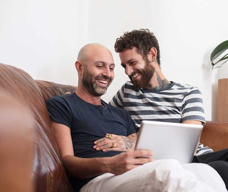 Männliches Paar kuschelt auf dem Sofa, lachen und schauen etwas auf einem Tablet