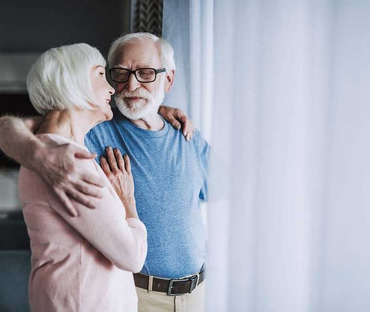 Älteres Paar steht am Fenster, umarmt sich und schaut sich in die Augen