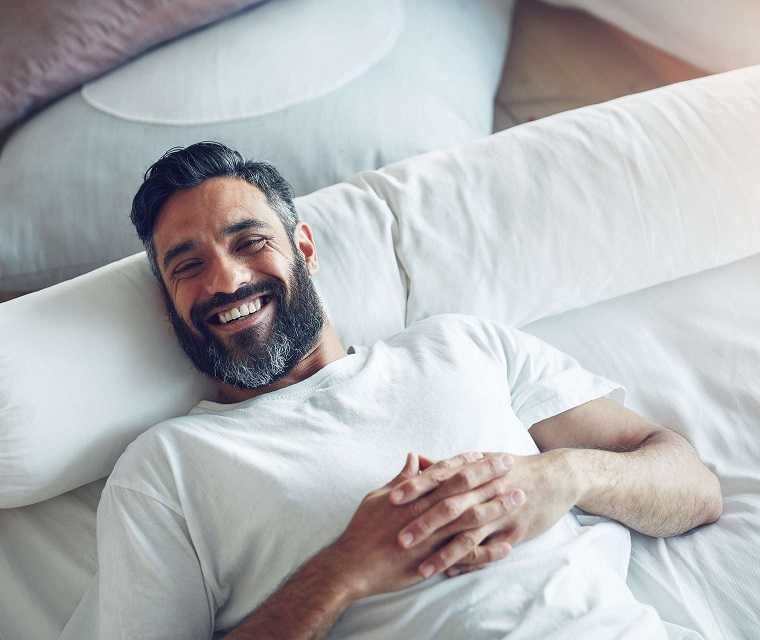 Mann mit Vollbart liegt im Bett und lacht in die Kamera Mann mit Vollbart liegt im Bett und lacht in die Kamera