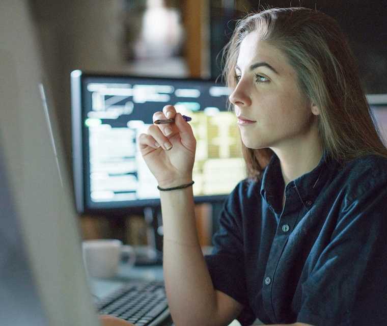 Junge Frau schaut mit einem Stift in der Hand auf einen Bildschirm und arbeitet