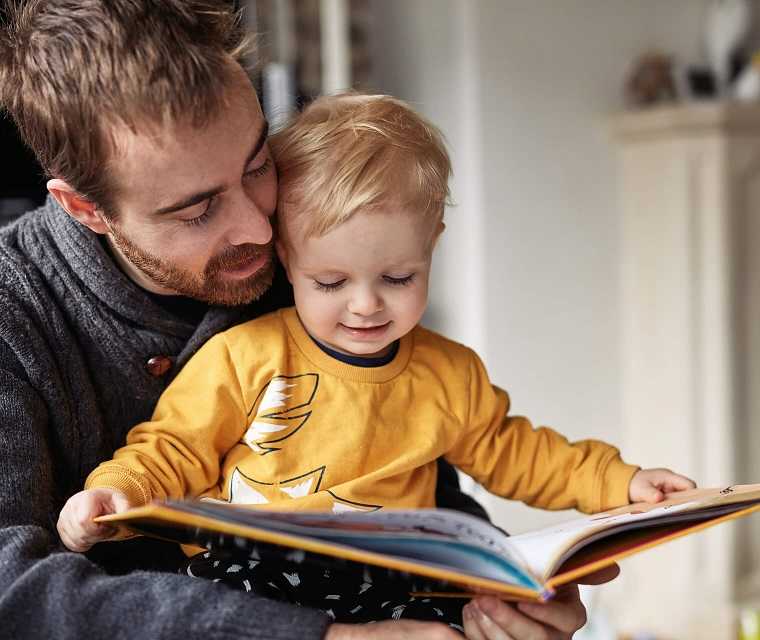 Vater hat ein Kind auf dem Schoß und sie schauen ein Bilderbuch an Vater hat ein Kind auf dem Schoß und sie schauen ein Bilderbuch an