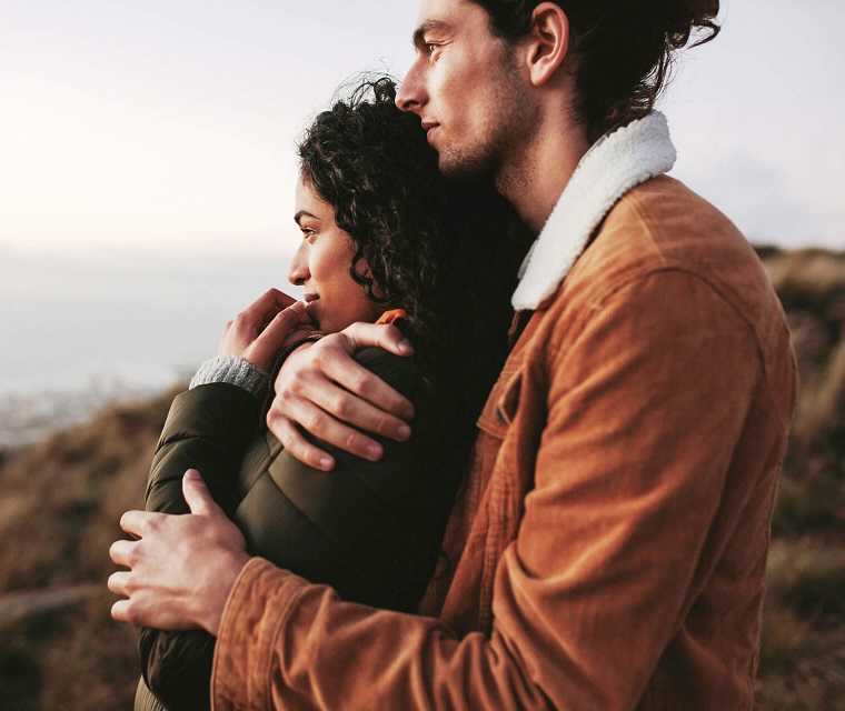 Junger Mann umarmt seine Freundin von hinten und beide schauen in die Ferne