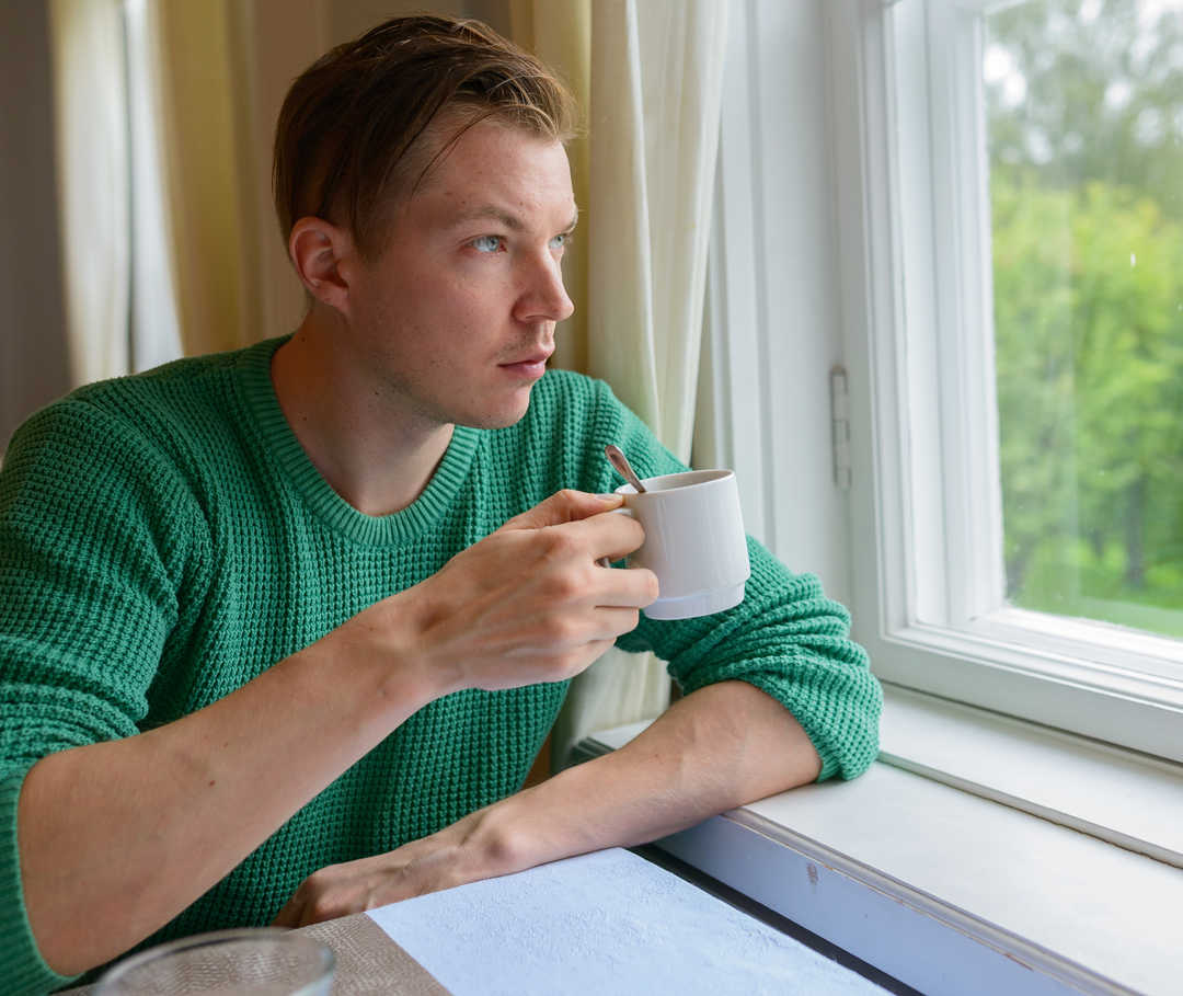 Ratgeber Rheuma: Ein Mann schaut mit einer Tasse in der Hand aus einem Fenster Ratgeber Rheuma: Ein Mann schaut mit einer Tasse in der Hand aus einem Fenster