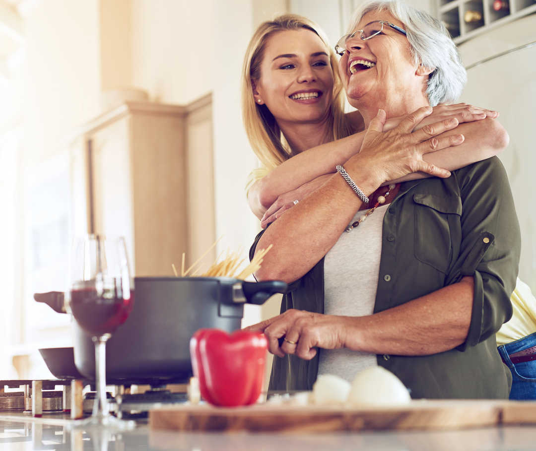 Ratgeber Rheuma: Eine erwachsene Tocher umarmt ihre Mutter, die am Herd steht
