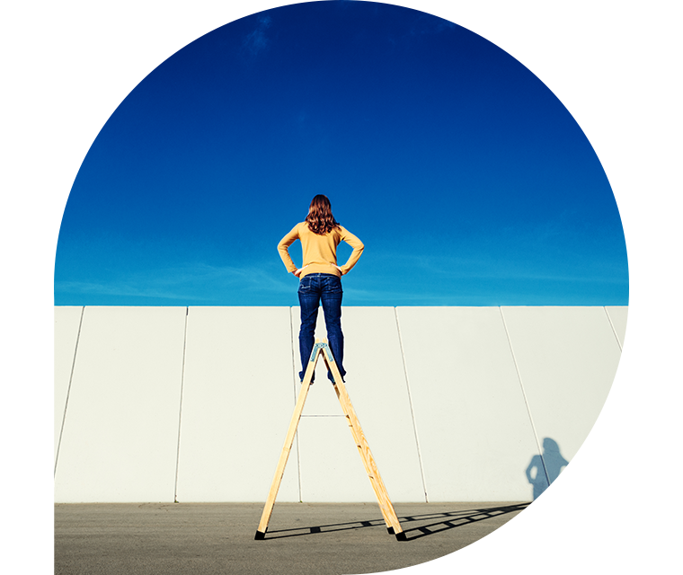 Ein Frau steht bei strahlend blauem Himmel auf einer Leiter und blickt über eine Mauer
