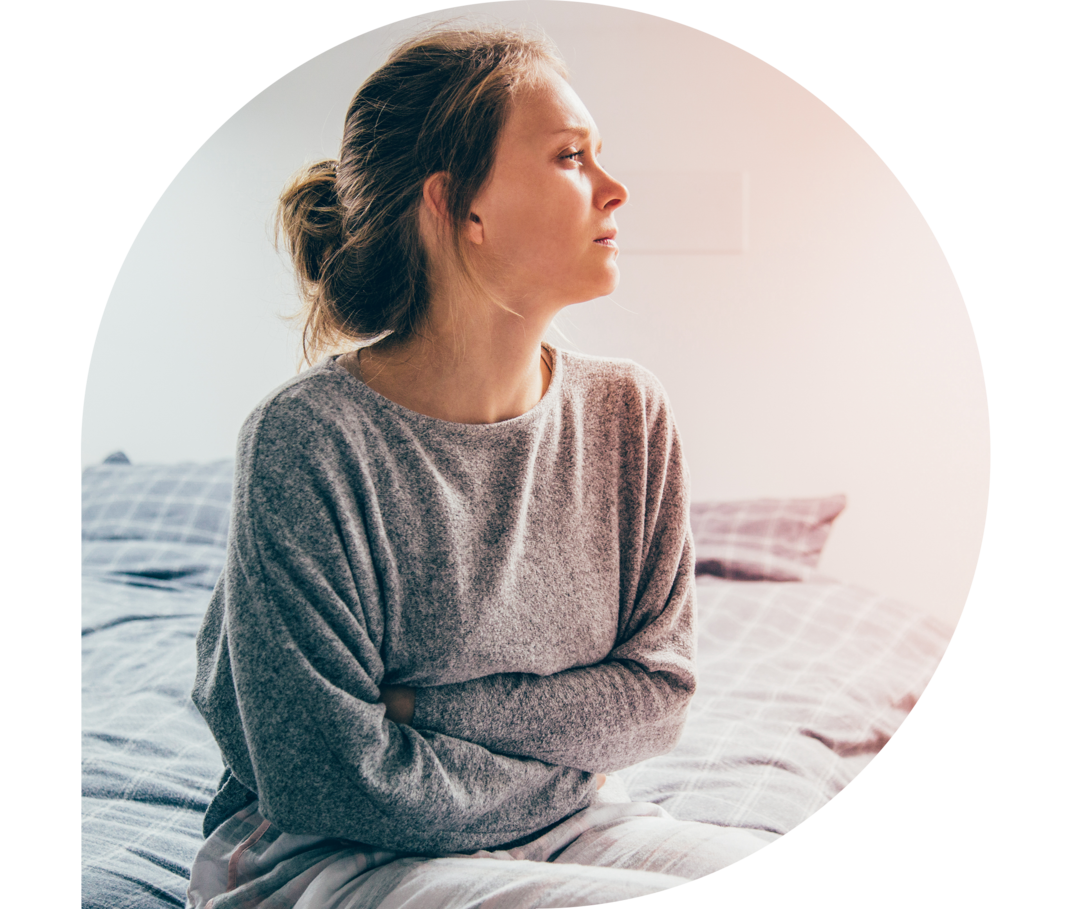 Eine Frau sitzt mit nachdenklichem Blick auf einem Bett und hält sich den Oberkörper.
