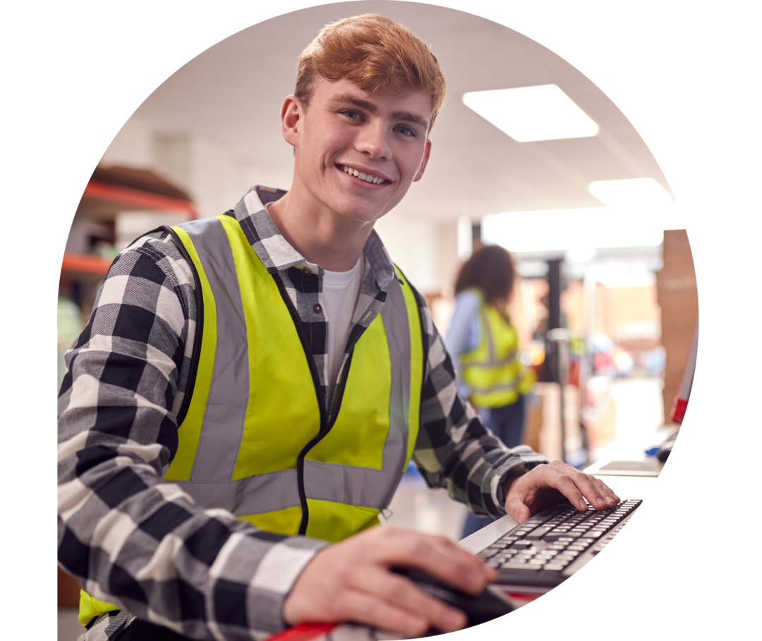 Ein junger Mann mit Arbeitsweste sitzt in einem Lager an einem Computerterminal