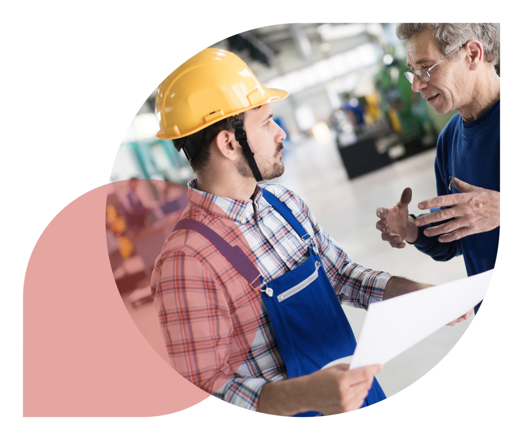 Ein Mann im Blaumann und mit Schutzhelm unterhält sich in einer Werkshalle mit einem Kollegen.