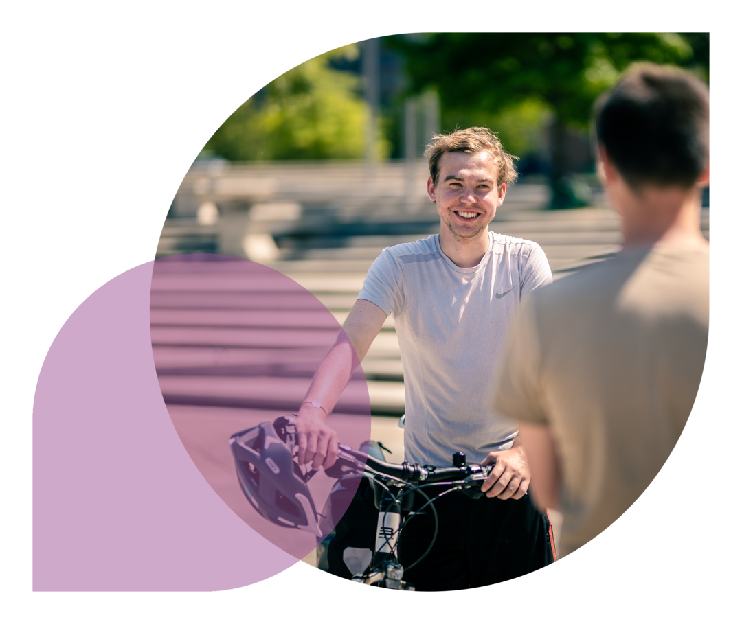 Phil Oliver Ladehof trägt ein weißes Shirt, hält ein Fahrrad fest und spricht mit einer Person, die von hinten zu sehen ist.