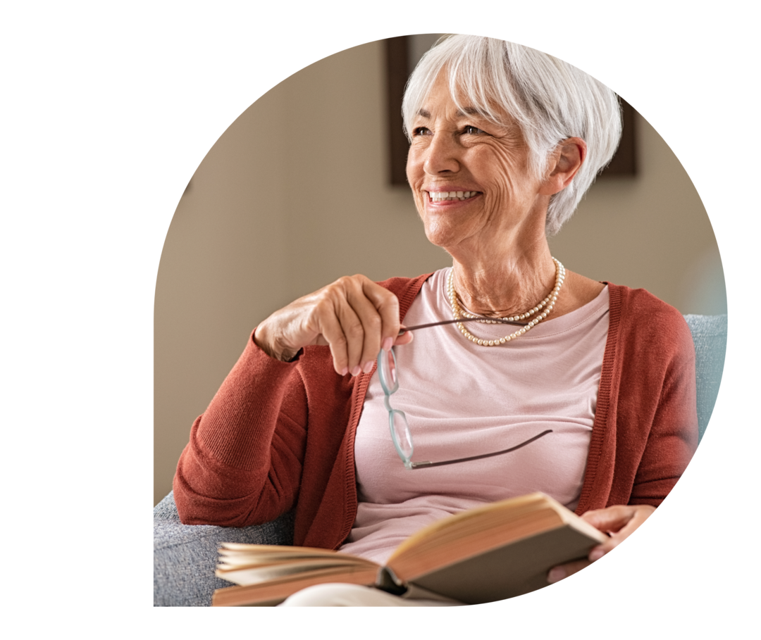 Eine grauhaarige Frau sitzt mit einem Buch in einem Sessel. In der rechten Hand hält sie eine Brille.