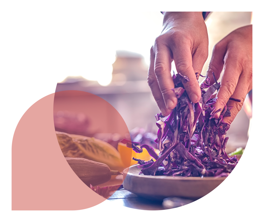 Zwei Hände einer Person, die geschnittenen Rotkohl auf einem Holzbrett anrichtet. Daneben liegen eine Gurke und Salatköpfe.
